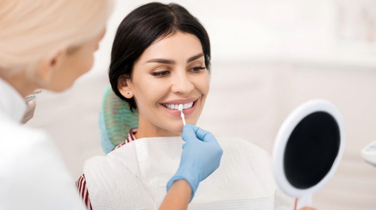 A woman at a dental clinic smiles while a dentist applies a whitening treatment to her teeth, holding a shade guide for comparison.